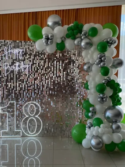 An 18th birthday with a green, white, and silver balloon arch against a sequin wall with neon signs.