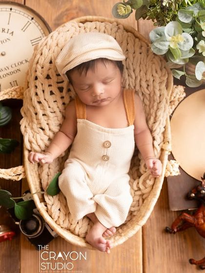 A newborn dressed in a vintage-style outfit with a newsboy cap sleeps in a basket, surrounded by classic props like a clock and toy animals.