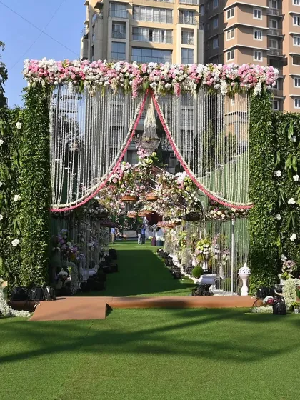 The grand entrance to an outdoor wedding ceremony, featuring a lush green walkway and a beautiful archway made of flowers and intricate metalwork.