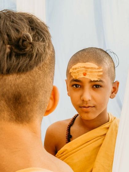A candid portrait of a young Batuk, his face marked with sandalwood paste, seen from behind another student. This shot captures the innocence and curiosity of youth in a deeply traditional setting.