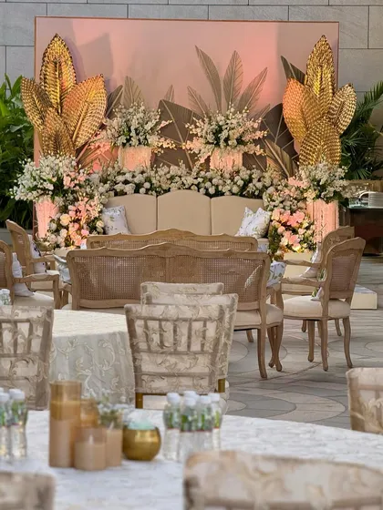 A beautiful stage setup combining white, gold, and green elements. The backdrop features a soft tropical print, flanked by large golden leaf sculptures and an abundance of white flowers. 🍃