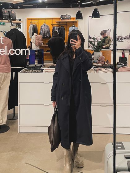 A mirror selfie taken in a retail store, showing a chic, all-black outfit with a long trench coat.