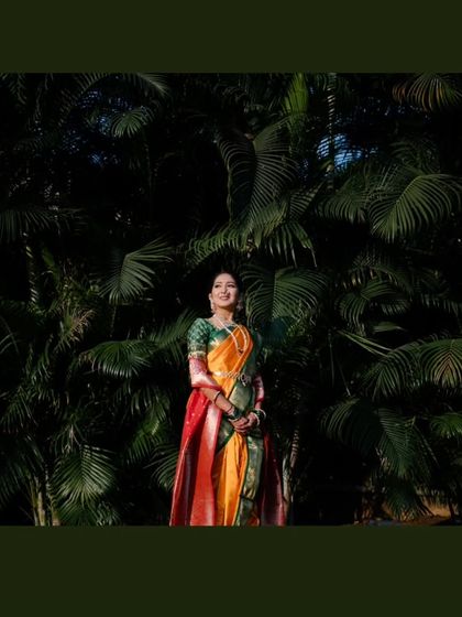 A stunning full-length portrait of the bride standing against a dark green, leafy backdrop, which makes her vibrant saree stand out.
