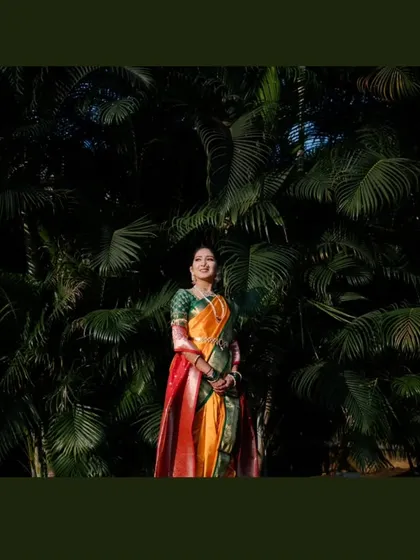 A stunning full-length portrait of the bride standing against a dark green, leafy backdrop, which makes her vibrant saree stand out.