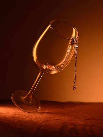 A delicate chain necklace draped inside a wine glass. The warm, amber lighting and elegant composition create a sense of sophistication and after-dark glamour.