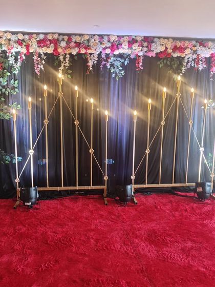 A rich and dramatic backdrop perfect for a Mehndi or Sangeet night. The dark curtain is framed by a dense floral border, with elegant candelabras and floor-level uplights adding a warm, golden glow to the stage area.