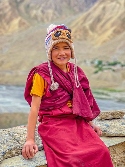 A smiling young monk in a playful panda hat sits on a wall in Spiti. This photo captures the lighter side of monastic life and the universal joy of childhood.