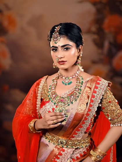A poised and elegant bridal portrait. The combination of the rich saree, heavy jewelry, and the artistic floral backdrop creates a look of timeless beauty.