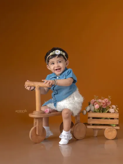 A sweet baby girl in a denim outfit takes a ride on a wooden tricycle. The simple, warm background and her happy smile make this a delightful and timeless photo.