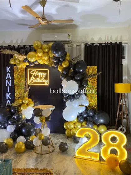 A chic 28th birthday party with a black and gold theme. The decor includes a balloon arch against a dark backdrop with a gold shimmer panel, a neon sign, and a light-up number.