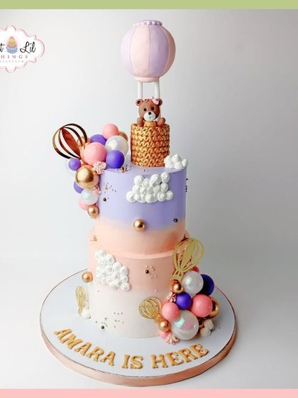"Amara is here!" A dreamy welcome cake for a new baby, featuring a two-tiered design with hot air balloons, clouds, and a teddy bear.