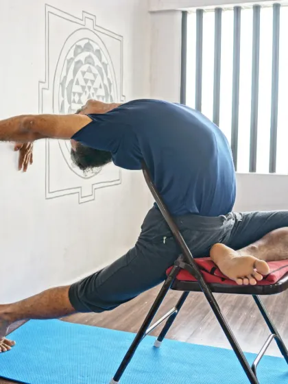 A variation of Eka Pada Rajakapotasana (Pigeon Pose) on a chair, using the wall for support. This modification allows for a deep hip and quad stretch while keeping the pose stable.