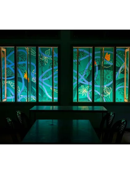 The view from inside the cafe at night. The hand-painted louvers, featuring cool blue and green tones on the interior side, become a beautiful, illuminated piece of art, creating a magical atmosphere for the children.