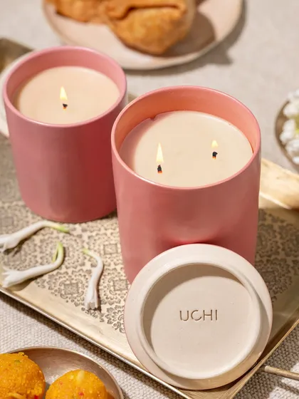 Two beautifully lit scented candles in pink ceramic jars, styled on a golden tray with Indian sweets and flowers. The warm glow of the flames creates a cozy and festive atmosphere, suggesting relaxation and cultural richness.
