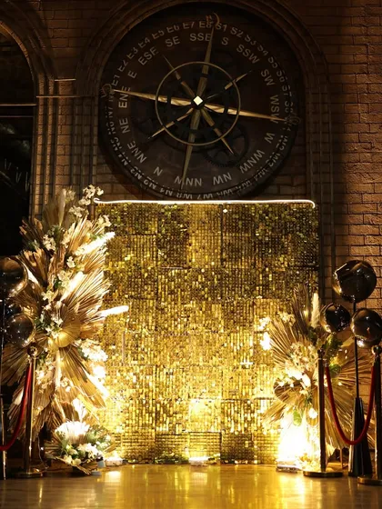 A glamorous gold shimmer wall serves as the perfect photo backdrop for a club event, complemented by boho-style dried floral arrangements.