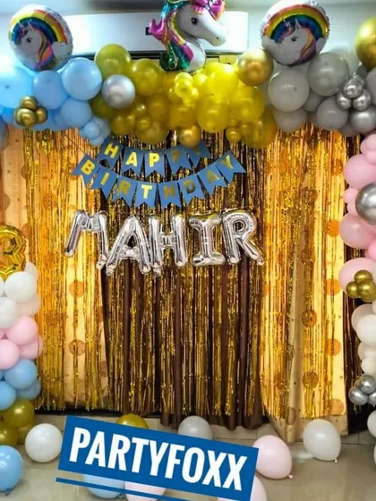A unicorn-themed birthday decoration for Mahir. This home setup includes a gold fringe backdrop and a colorful balloon arrangement with unicorn foil balloons.