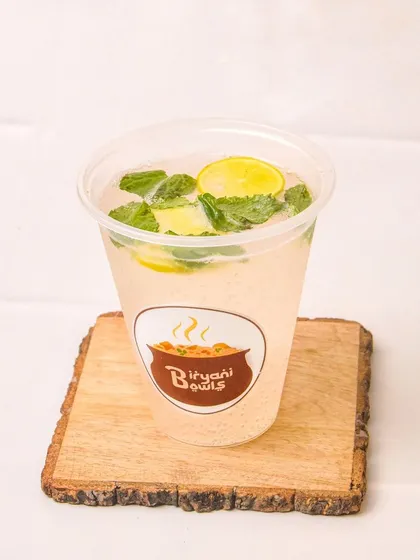 A glass of masala lemonade with mint and lemon slices, presented on a wooden coaster. The clean background makes this a crisp and refreshing beverage shot.