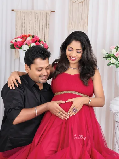 A joyful couple portrait in a studio setting. The husband makes a heart shape with his hands on his wife's bump, and her bright smile shows their shared happiness.
