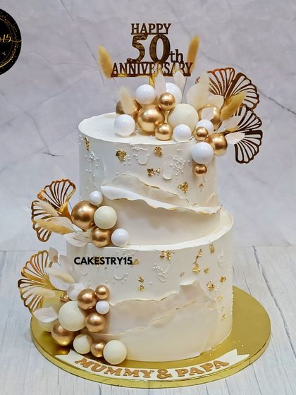 An elegant 5kg, two-tier chocolate cake for a 50th anniversary. The ivory-colored cake is decorated with white and gold decorative balls, golden ferns, and a "Mummy & Papa" nameplate.