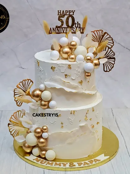 An elegant 5kg, two-tier chocolate cake for a 50th anniversary. The ivory-colored cake is decorated with white and gold decorative balls, golden ferns, and a "Mummy & Papa" nameplate.