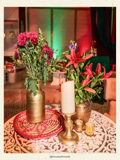 A close-up of a table centerpiece with red and orange flowers in a gold vase, set on a carved white table, creating a rich and vibrant look.