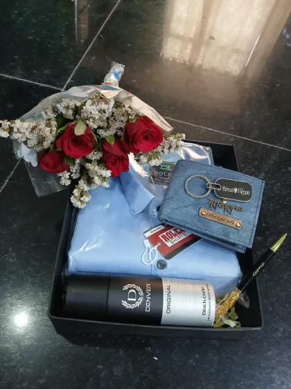 A beautifully arranged gift hamper with a bouquet of red roses, a personalized denim wallet and keychain, a shirt, a pen, and deodorant.