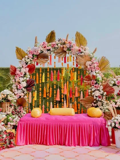 A beautiful Mehendi seating arrangement combining a vibrant pink couch with a floral arch of roses and dried palms. This boho-luxe design is perfect for a stylish and comfortable bridal seating area.