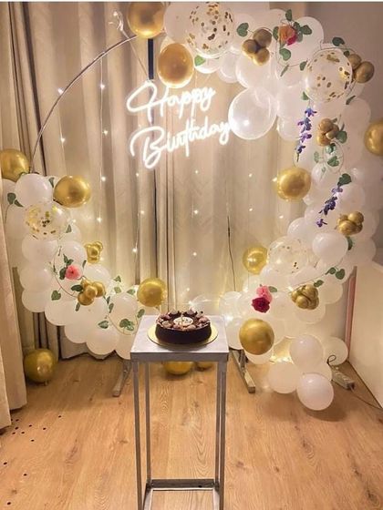 A simple yet beautiful white and gold balloon ring with a cake stand. This setup is perfect for an intimate celebration, with confetti balloons and a 'Happy Birthday' neon sign.