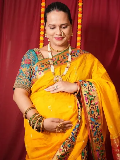 A classic pose for a maternity photoshoot, this mom-to-be is wearing a vibrant yellow Nauvari saree. The studio setup includes traditional elements like marigold flower strings.