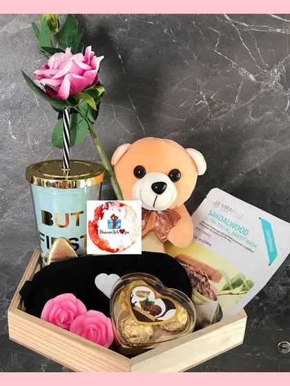 A curated self-care hamper perfect for Women's Day or as a gift for her. This wooden tray arrangement includes a cute teddy bear, a "But First..." tumbler, a sandalwood face mask, an eye mask, and chocolates.