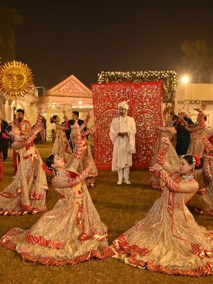 A full view of the grand groom entry, with dancers and prop holders creating a majestic welcome. The scene is set against a beautifully carved red backdrop, adding to the opulence.