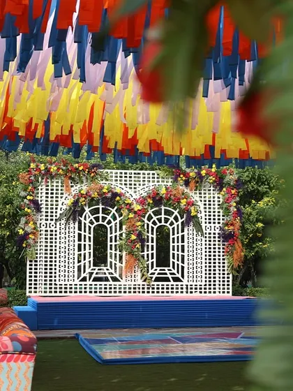 A colorful Haldi setup at the Taj Palace, New Delhi. The decor features a ceiling of multi-colored ribbons and a stage with a unique white lattice backdrop.
