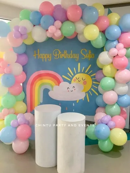 A pastel rainbow themed birthday party. The decoration features a cute sun and cloud backdrop framed by a soft-colored balloon arch.