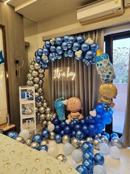 A stylish and modern "It's a Boy" welcome home decoration. This setup features a half-moon balloon arch in metallic blue and silver, a neon sign, and cute props like baby foil balloons and transparent 'BABY' blocks.
