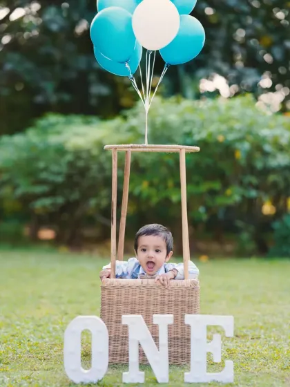 An adorable shot of a one-year-old peeking out of a hot-air balloon basket during his outdoor birthday shoot. The 'ONE' sign clearly marks the special occasion.
