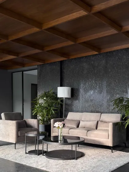 A formal waiting lounge that balances texture and tone. The area is defined by a dark, textured granite feature wall and a warm, wooden coffered ceiling, furnished with minimalist sofas and tables to create a sophisticated yet comfortable atmosphere.
