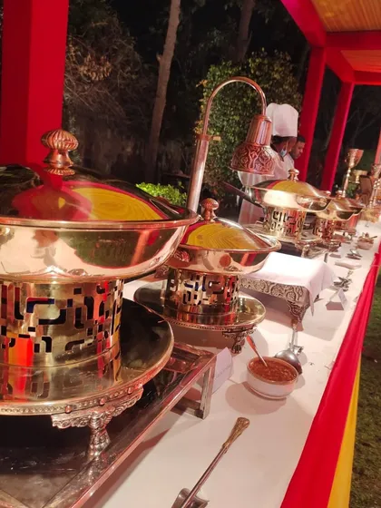 A close-up of the traditional brass and copper serving dishes, known as 'chafing dishes' or 'bain-maries', keeping the food warm. This setup is part of a wedding catering service in a colorful outdoor tent.