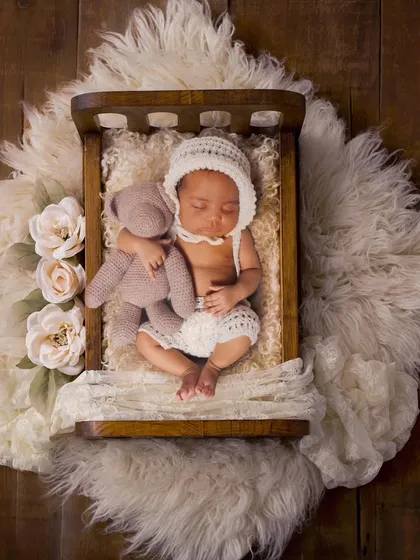 A 20-day-old baby sleeping in a tiny wooden bed with a teddy bear, set on a large fur rug.