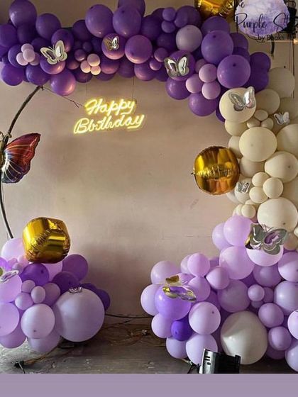 A stylish butterfly-themed balloon arrangement in purple and white, with a neon 'Happy Birthday' sign and large butterfly props.