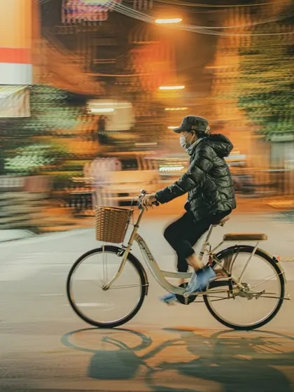 A person wearing a mask and hat rides a bicycle at night in Hanoi, captured with a panning motion blur technique that emphasizes speed.