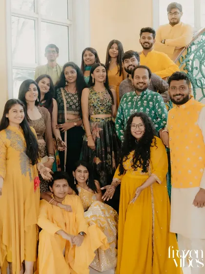 A large group of friends and family in vibrant yellow and green traditional wear pose together on a staircase. This photo captures the festive atmosphere and collective joy of a Haldi or Mehendi ceremony.