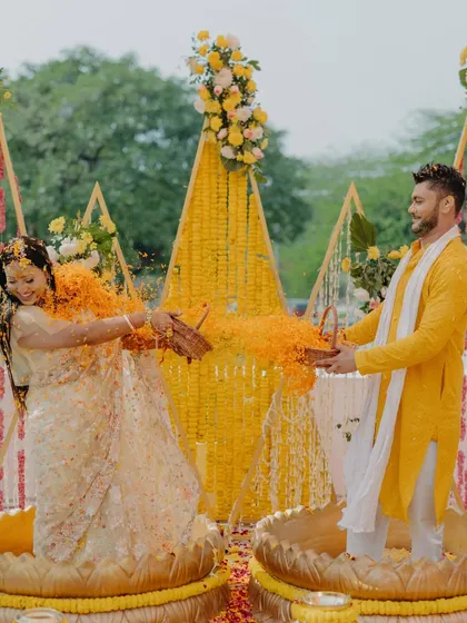 Capturing a moment of pure joy during a Haldi ceremony. The decor features triangular frames draped in marigolds, with the couple seated in large brass 'urli' bowls for the rituals, making the experience truly unique and memorable.