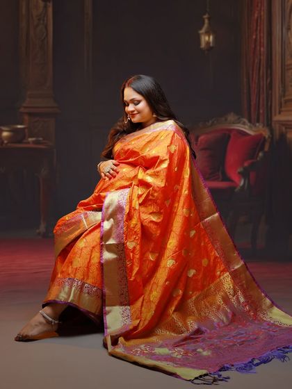 A serene seated portrait of the mother-to-be. The flowing orange saree and the dimly lit, classic interior backdrop create a moody and artistic feel.