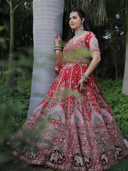 A candid shot of the bride in the traditional red lehenga, set against lush greenery. The natural light brings out the rich colors and detailed silver work.
