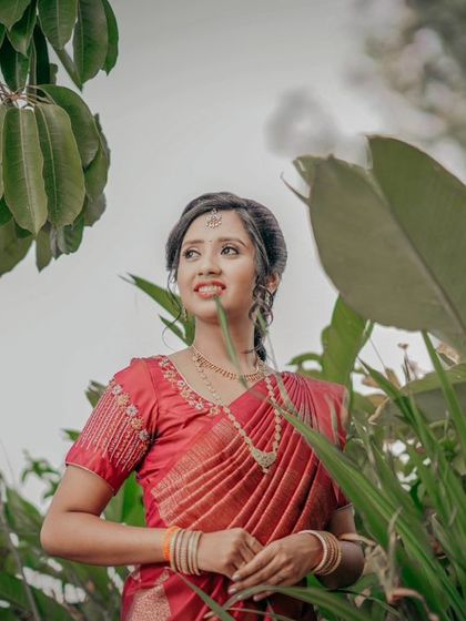 A bride in a red saree, surrounded by lush greenery, looking off to the side with a gentle smile.
