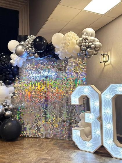 A sophisticated 30th birthday setup with a silver shimmer wall, a black and silver balloon arch, and large, bright marquee numbers.