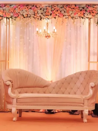 An elegant chaise lounge sits before a backdrop of sheer curtains and a crystal chandelier, framed by a delicate arrangement of peach and white flowers.