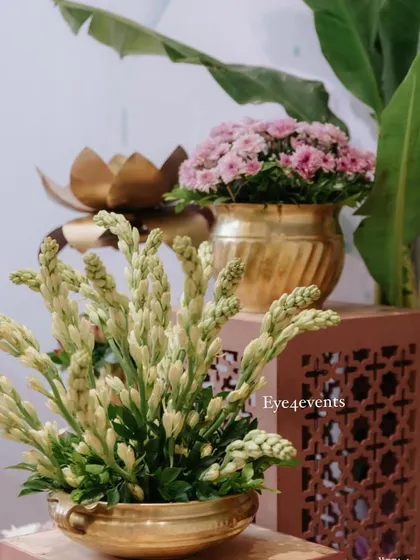This side decor arrangement combines traditional and modern elements. It features fresh tuberoses and pink flowers in elegant brass pots, placed on decorative stands, perfect for an engagement party or festive gathering.
