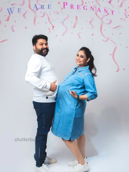 A fun and casual pregnancy announcement photo. The couple stands back-to-back with thumbs up, pointing to their bellies under a "We Are Pregnant" banner, sharing a laugh.