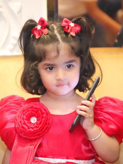 A little girl in a bright red dress with puffy sleeves holds a pen, looking thoughtfully at the camera. We capture these quirky and cute moments.